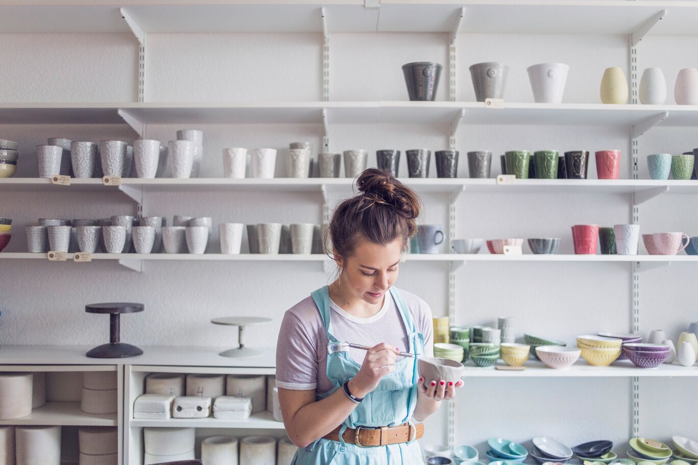 young-female-potter-using-hand-tool-on-vase-while-standing-against-shelves-at-store