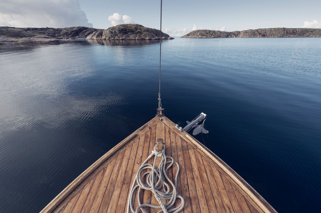 Du ser fören på en segelbåt i trä, du navigerar i ett kav lugnt skärgårdslandskap.