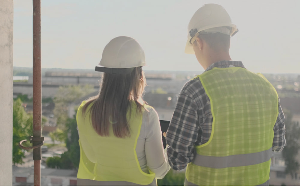 A man and a woman wearing hard hats and safety vests, looking out a window.