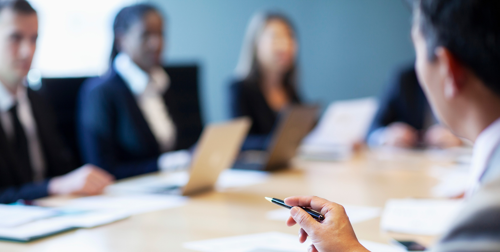 A group of business professionals in formal attire sit around a conference table during a meeting. Several laptops and documents are on the table, and one person in the foreground holds a pen while speaking. The background is softly blurred, keeping the focus on the discussion atmosphere