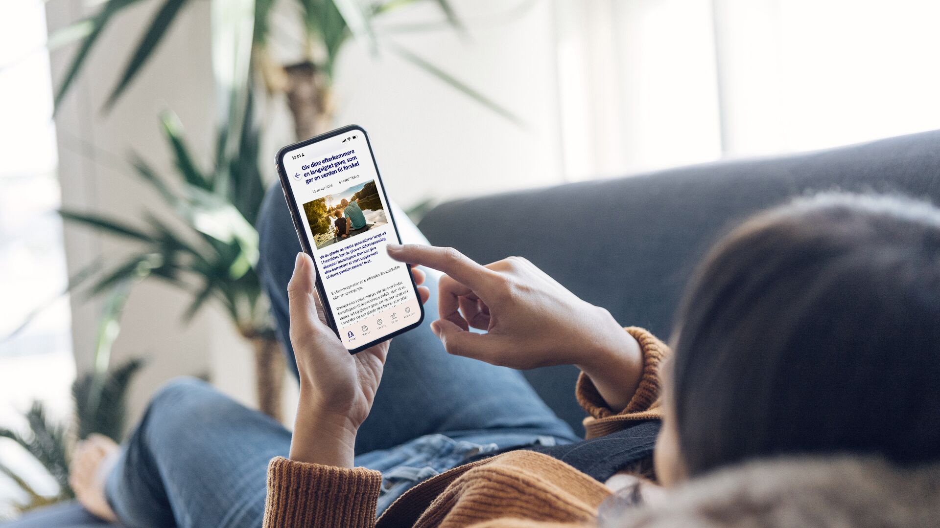 Foto af en person, der ligger afslappet i en sofa og holder en smartphone, hvor Nordea-appen MyFunds er åben på skærmen og viser en artikel om investeringer og opsparing.