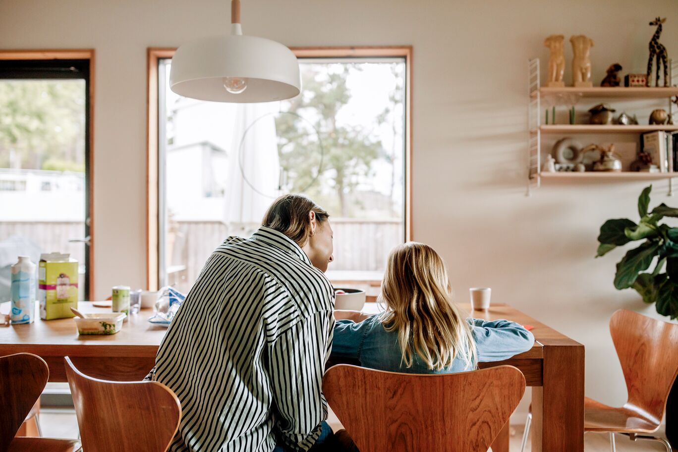 Adult and child sitting together at a wooden dining table, seen from behind, in a bright home with shelves and a pendant light
