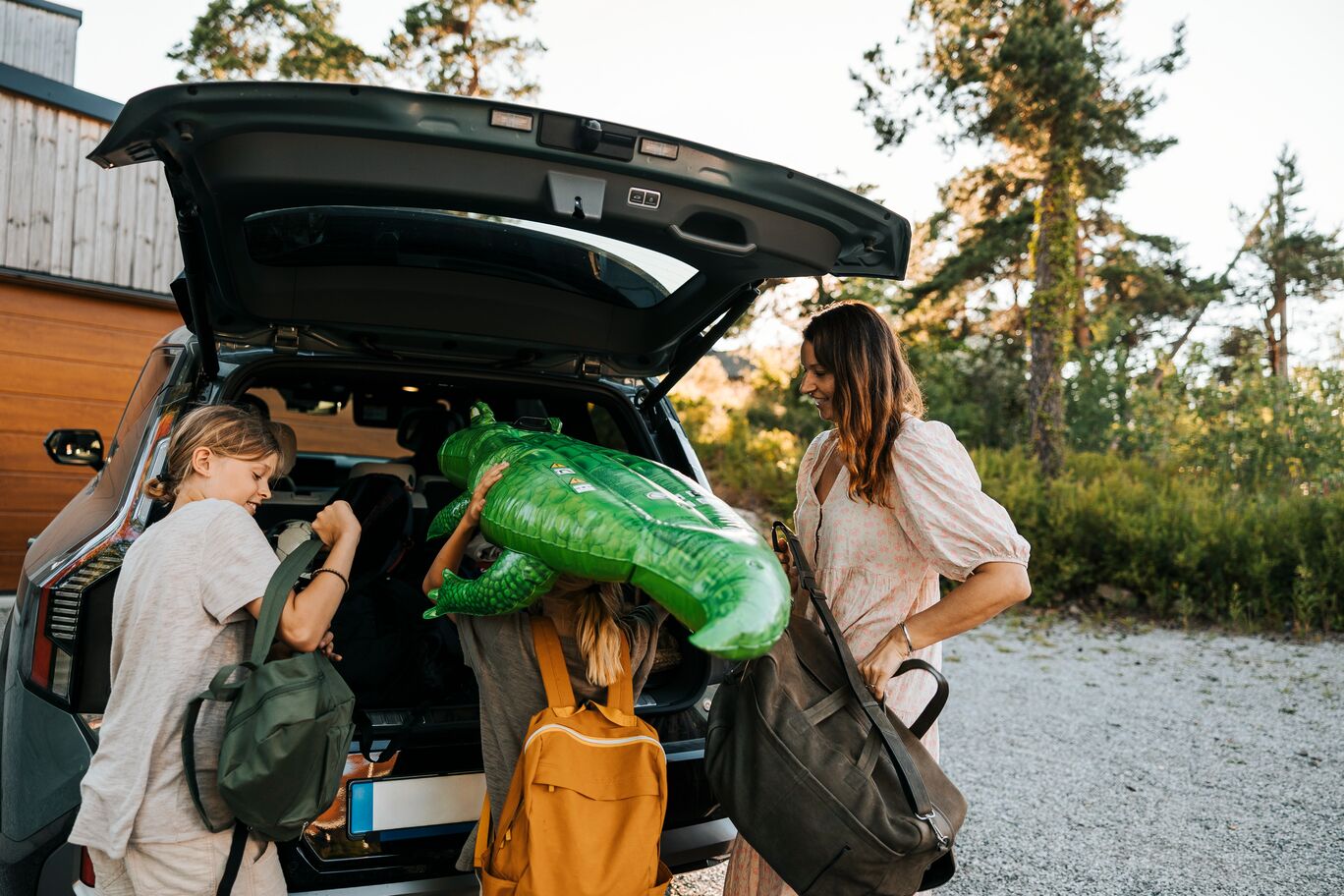 A woman and two children loading bags and an inflatable crocodile into the boot of a car, ready for a summer trip.