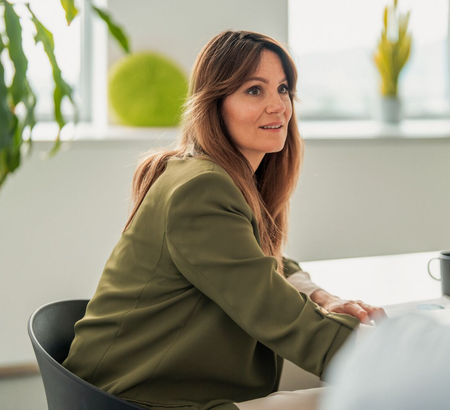 Woman in a green blazer sitting at a meeting table, listening attentively in a bright office with plants.