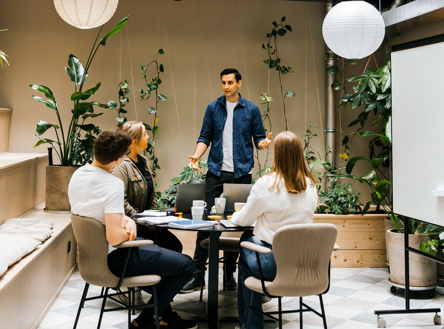A man presenting to three seated colleagues around a table in a bright, plant-filled office space with large white pendant lights.