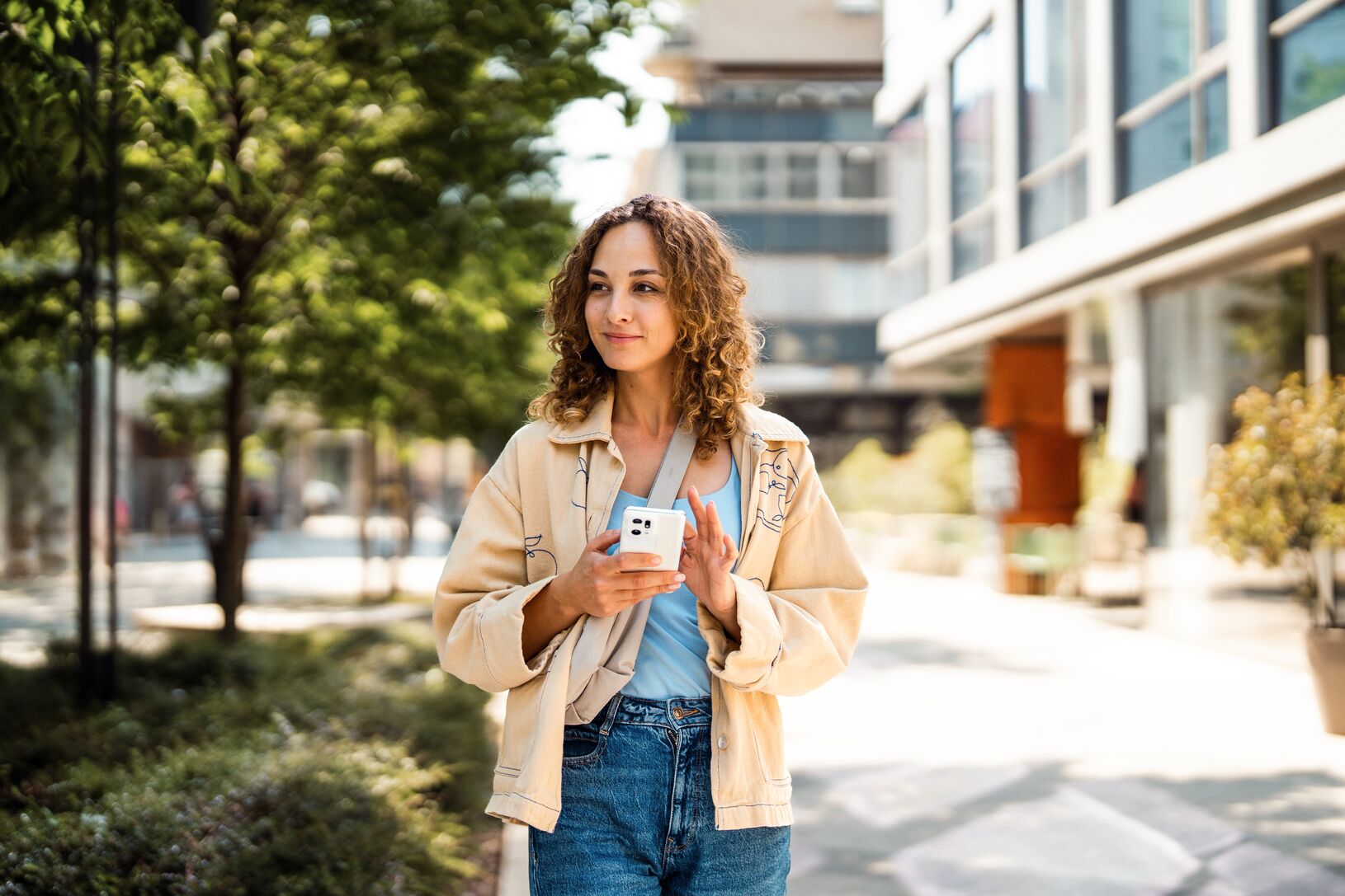 Young woman with curly hair holding a phone, walking along a sunny urban street lined with trees and shops.