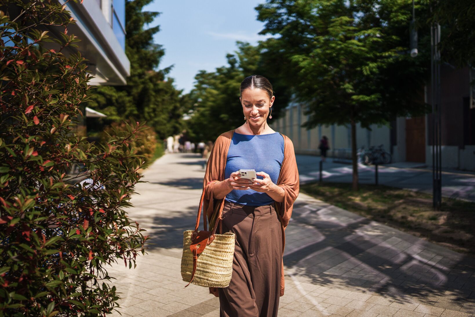 Woman walking along a sunny tree-lined path, smiling at her phone while carrying a straw basket bag.