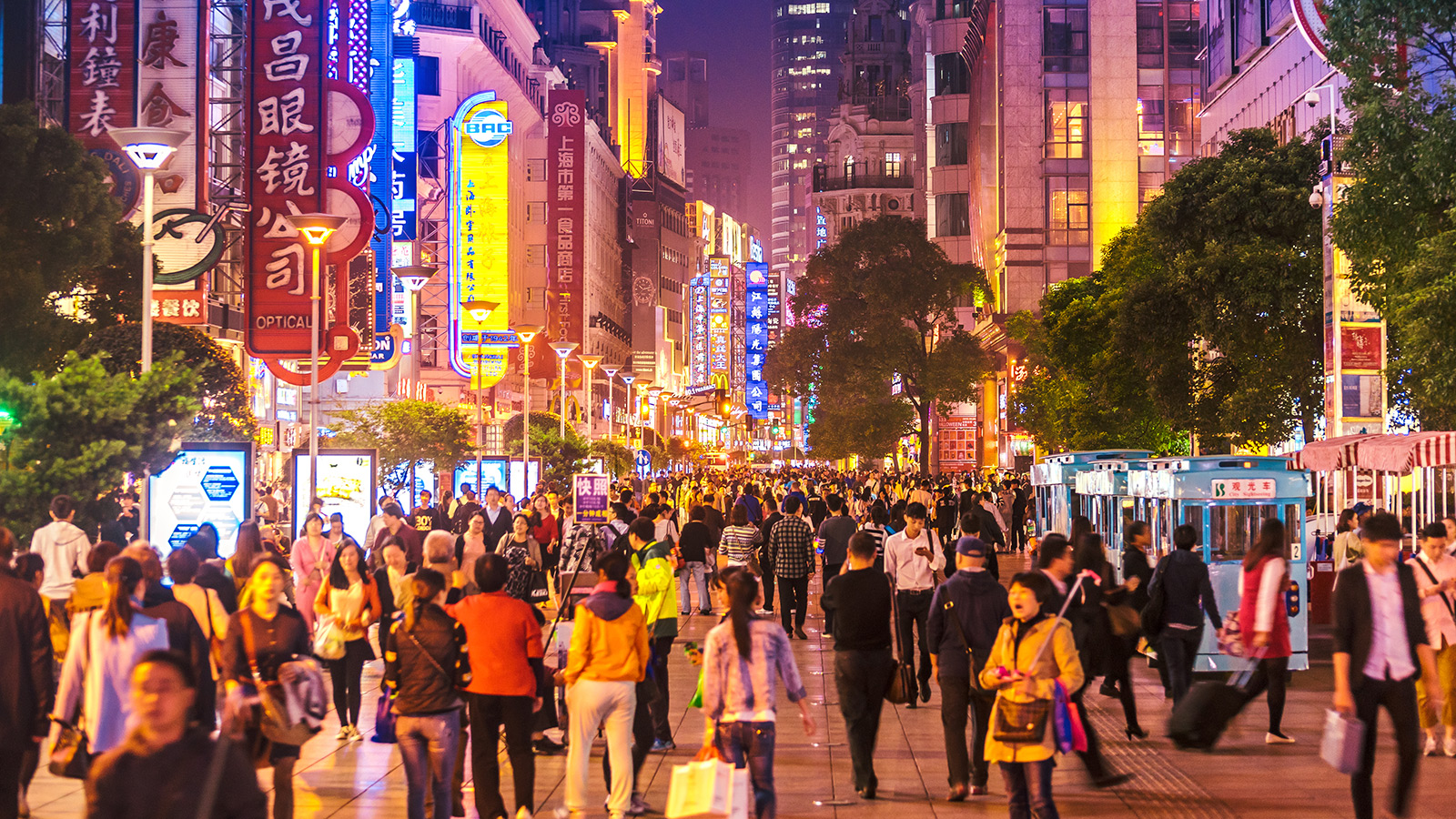 Crowds of people walking along a busy illuminated shopping street at night in a Chinese city, with colourful neon signs lining the buildings.