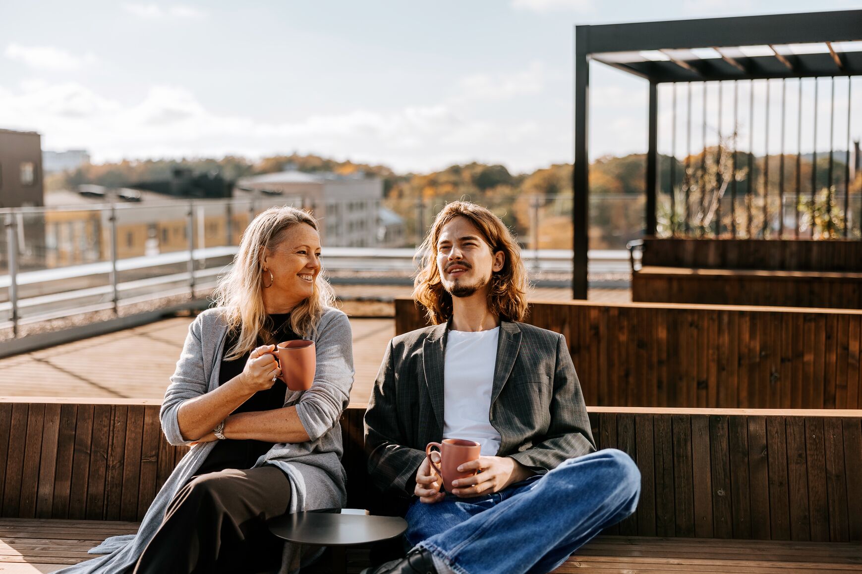 Two colleagues having a relaxed conversation over coffee on a rooftop terrace on a sunny autumn day.