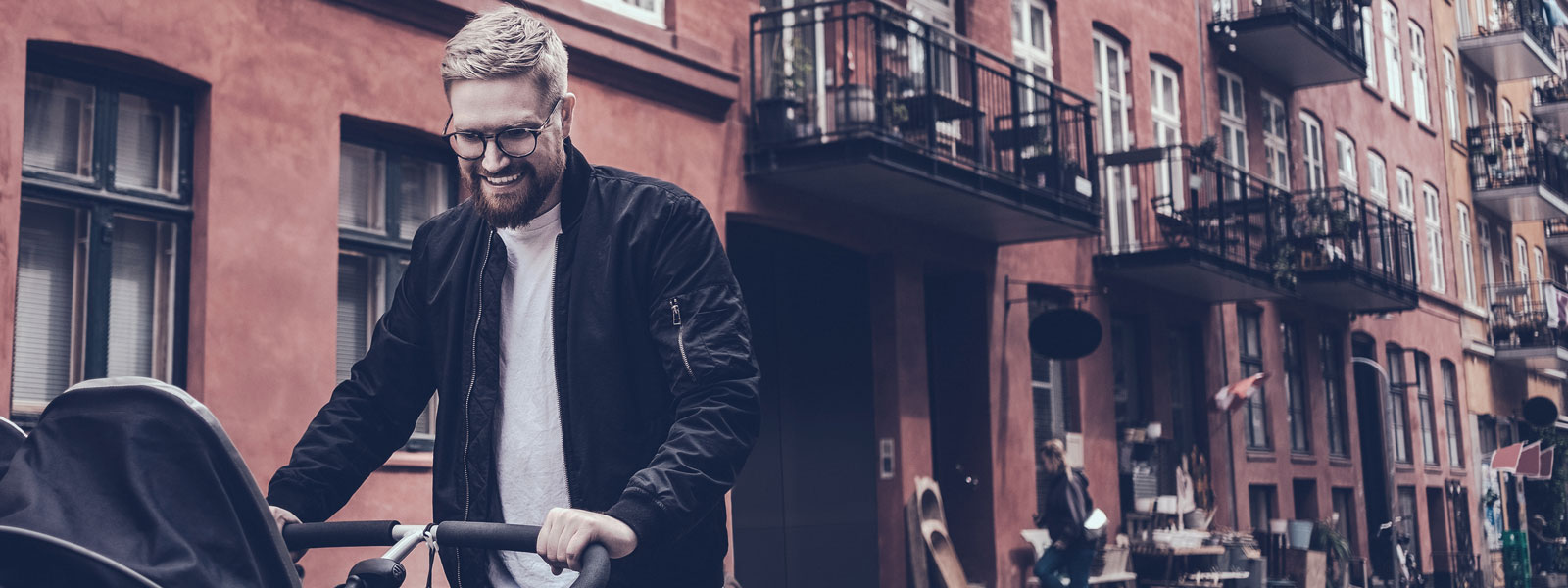 Smiling bearded man with glasses pushing a pram along a city street lined with red brick apartment buildings.