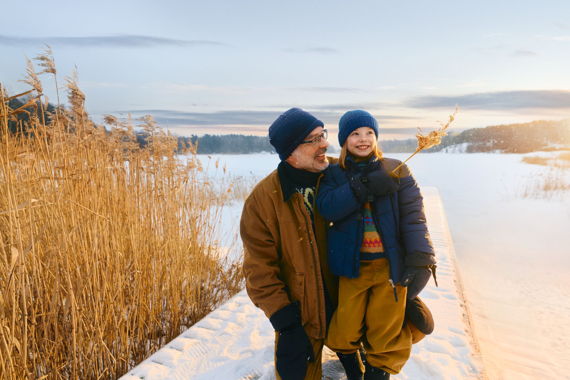 A man and a smiling girl in winter clothes standing together on a snowy lakeside path at sunset, surrounded by golden reeds.