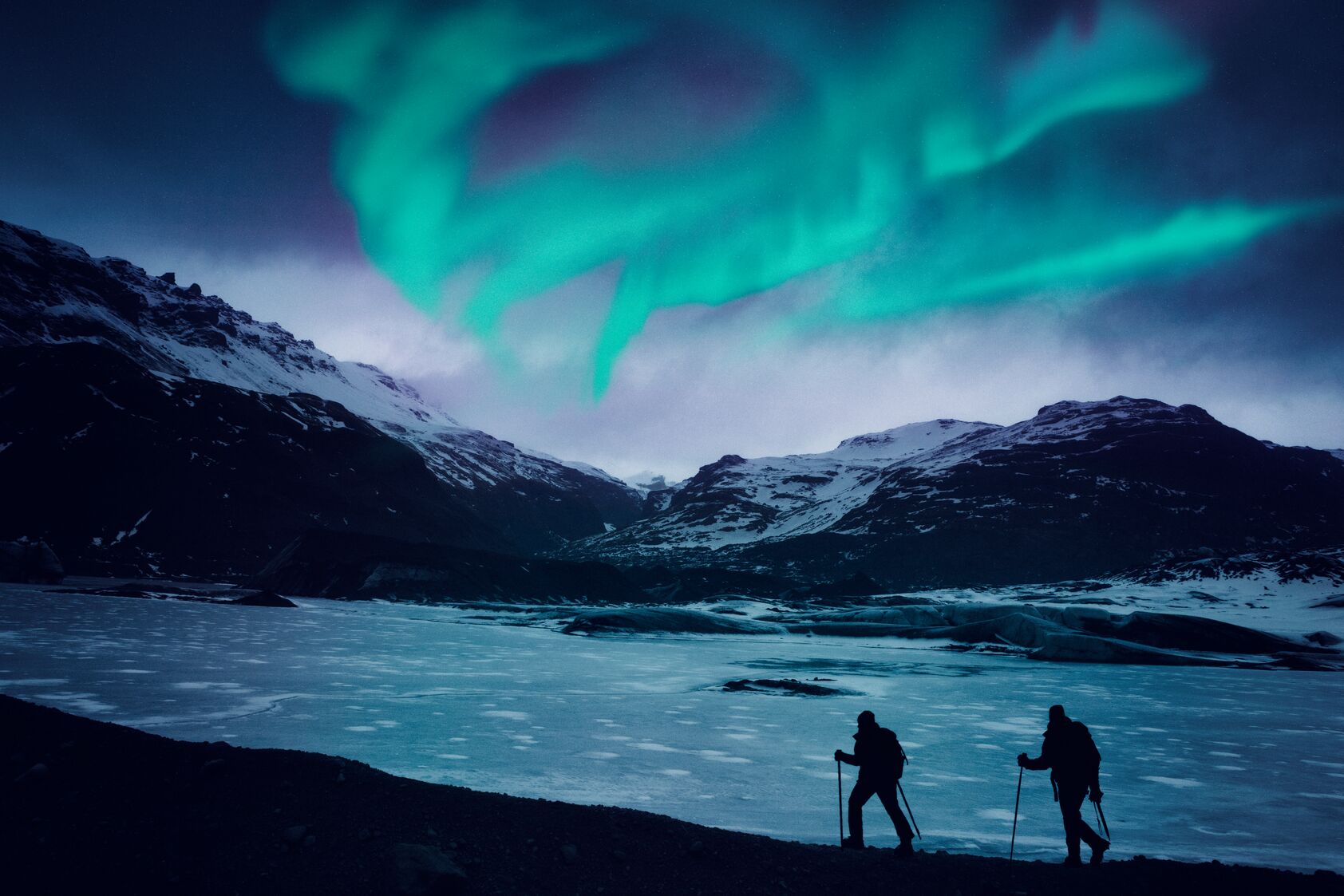 Two hikers with trekking poles silhouetted against a frozen glacial landscape beneath a vivid green northern lights display.
