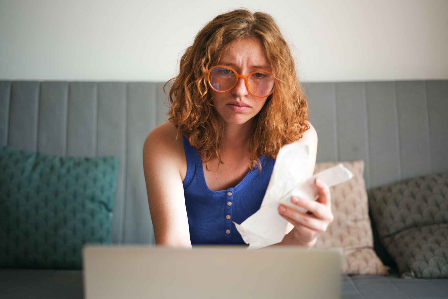 Woman with curly red hair and orange glasses looking concerned at a receipt while sitting in front of a laptop on a sofa.