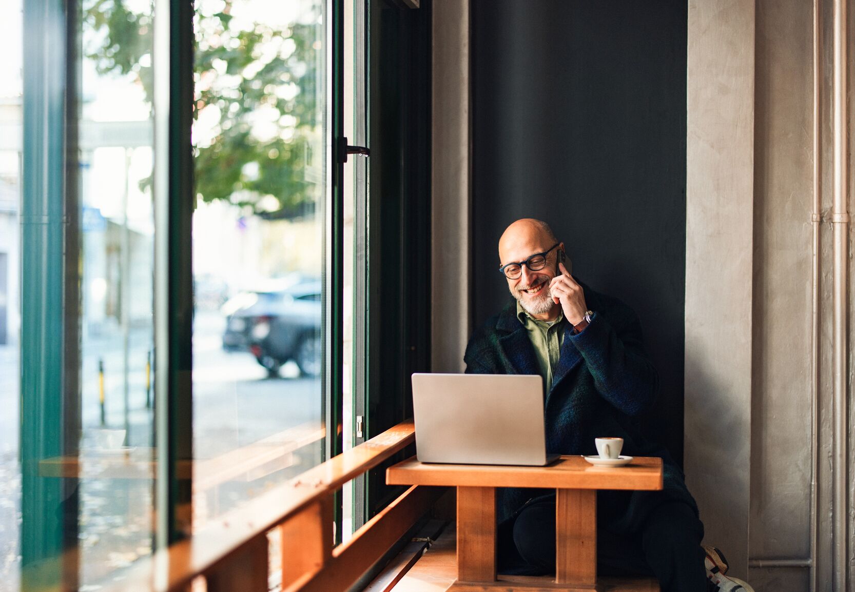 Smiling bald man with glasses talking on the phone at a café window seat, with a laptop and coffee cup on the table.