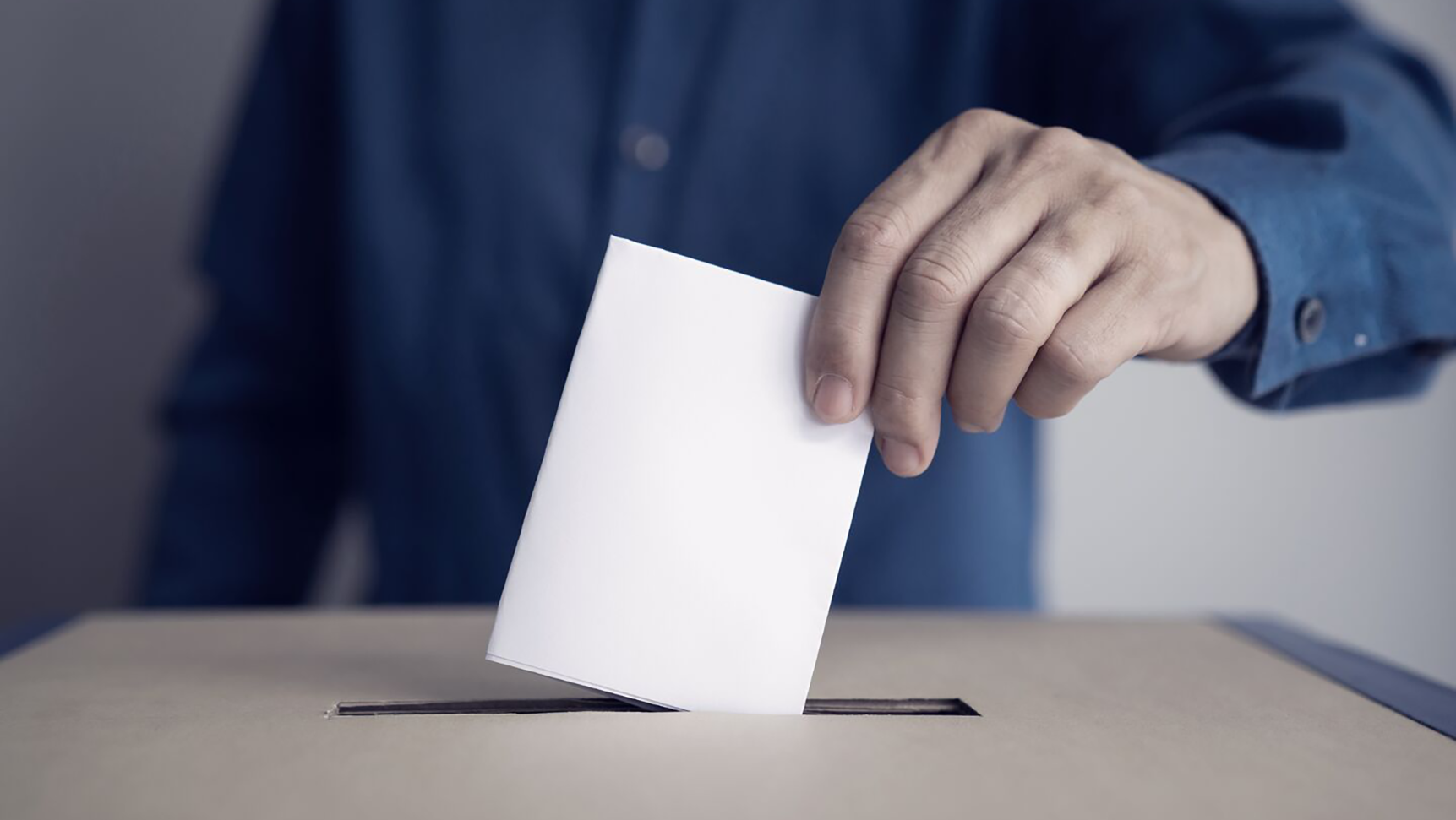 Foto af en person i blå skjorte, der lægger en hvid stemmeseddel i en stemmeboks af pap.