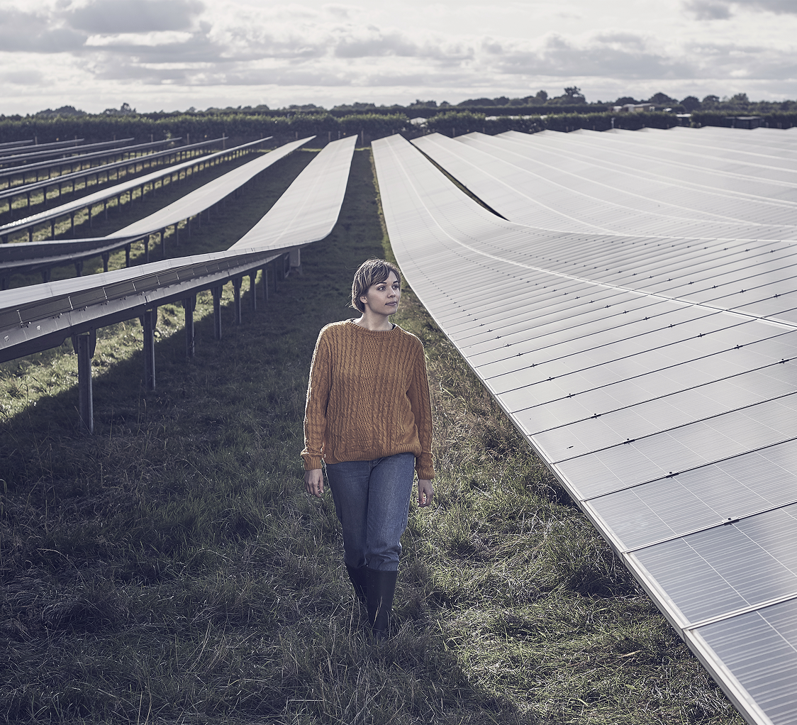 Woman in a knit sweater walking between rows of solar panels on a large solar farm under an overcast sky.