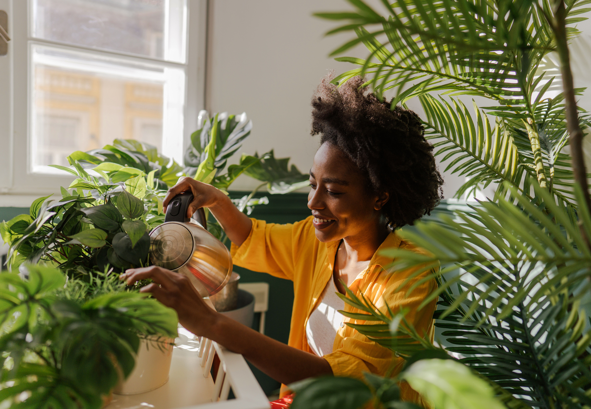 Smiling woman in a yellow shirt watering indoor plants at home, surrounded by lush greenery in a sunlit room.