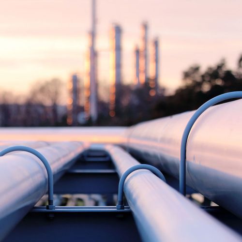 Et foto af store metalrør i et industriområde set i lav vinkel, hvor rørene strækker sig frem mod en sløret baggrund af fabriksstrukturer i solnedgangens bløde lys.