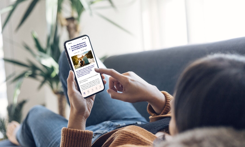 Foto af en person, der ligger afslappet i en sofa og holder en smartphone, hvor Nordea-appen MyFunds er åben på skærmen og viser en artikel om investeringer og opsparing.