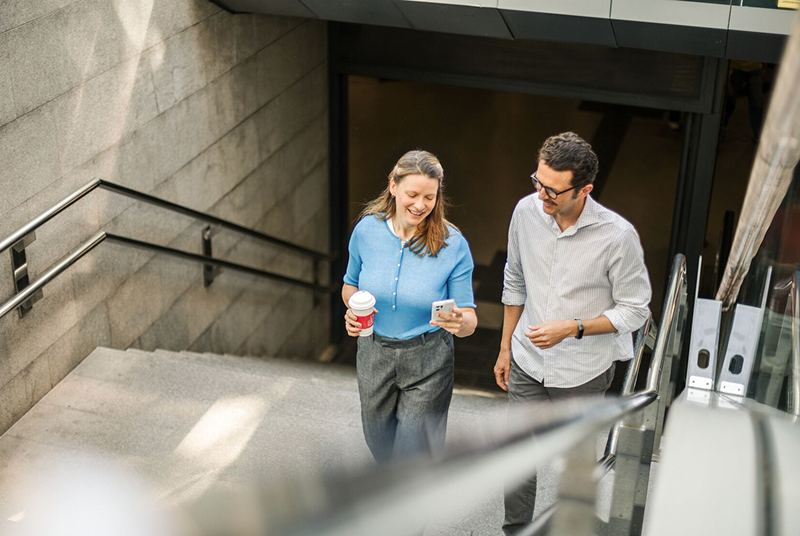 Two colleagues walking up stairs together, smiling and looking at a phone. One is holding a coffee cup.