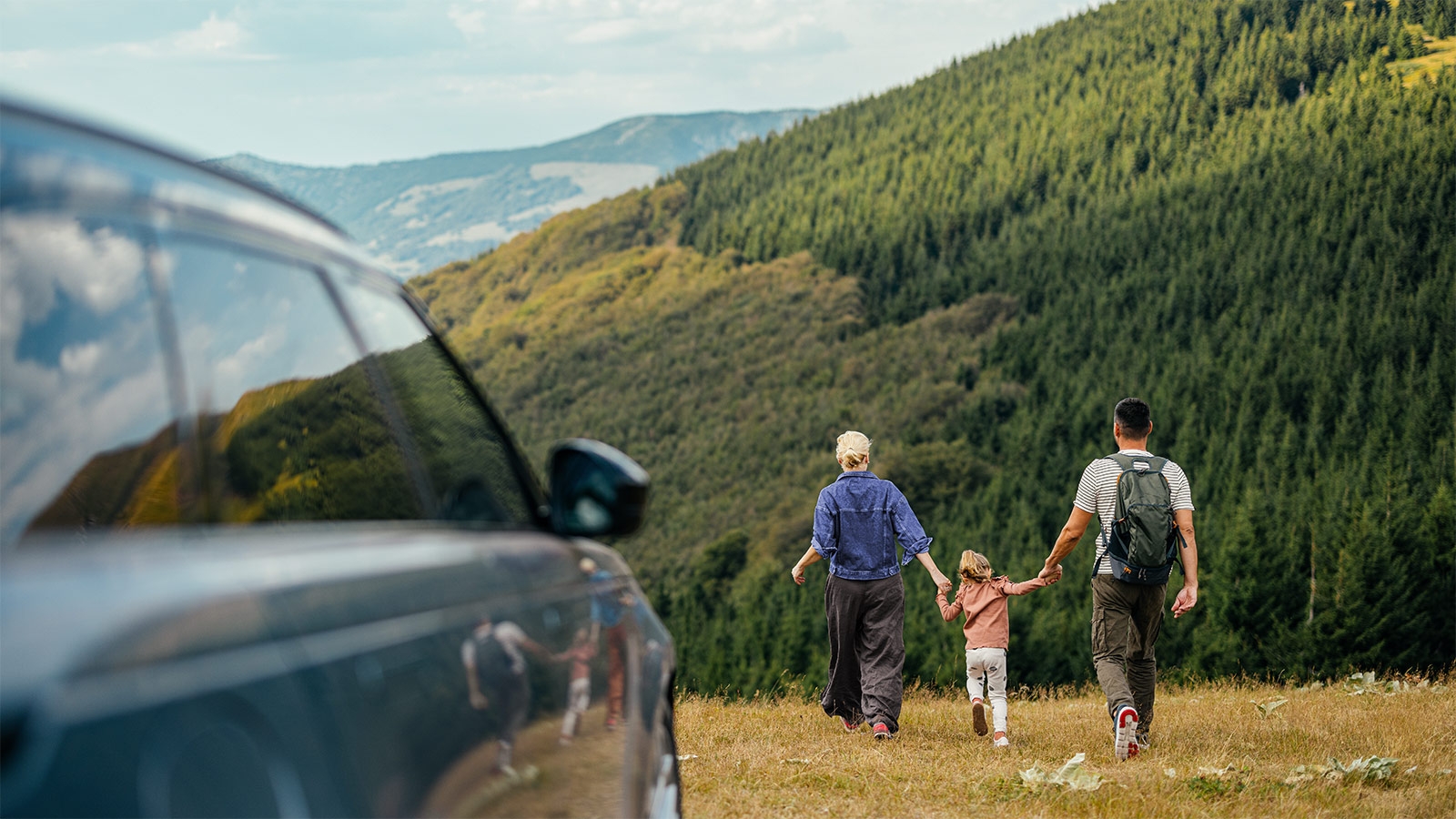 En familie nyder en campingtur i smukke bjergomgivelser, som fanger essensen af eventyr og samvær. Scenen byder på frodige grønne bakker og en parkeret bil.