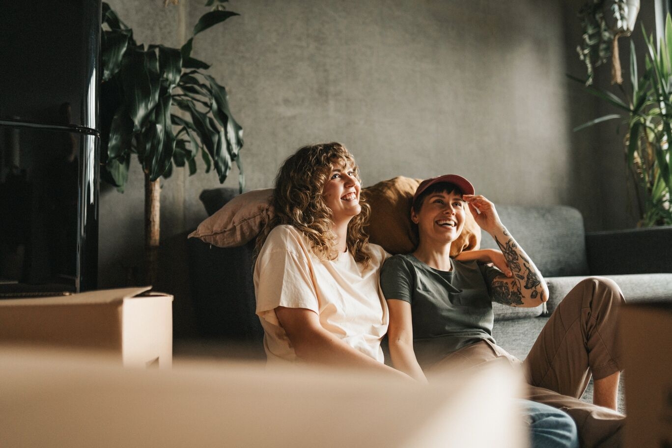Two women sitting on the floor laughing and looking up, relaxing together in a cosy living room with plants and a sofa.