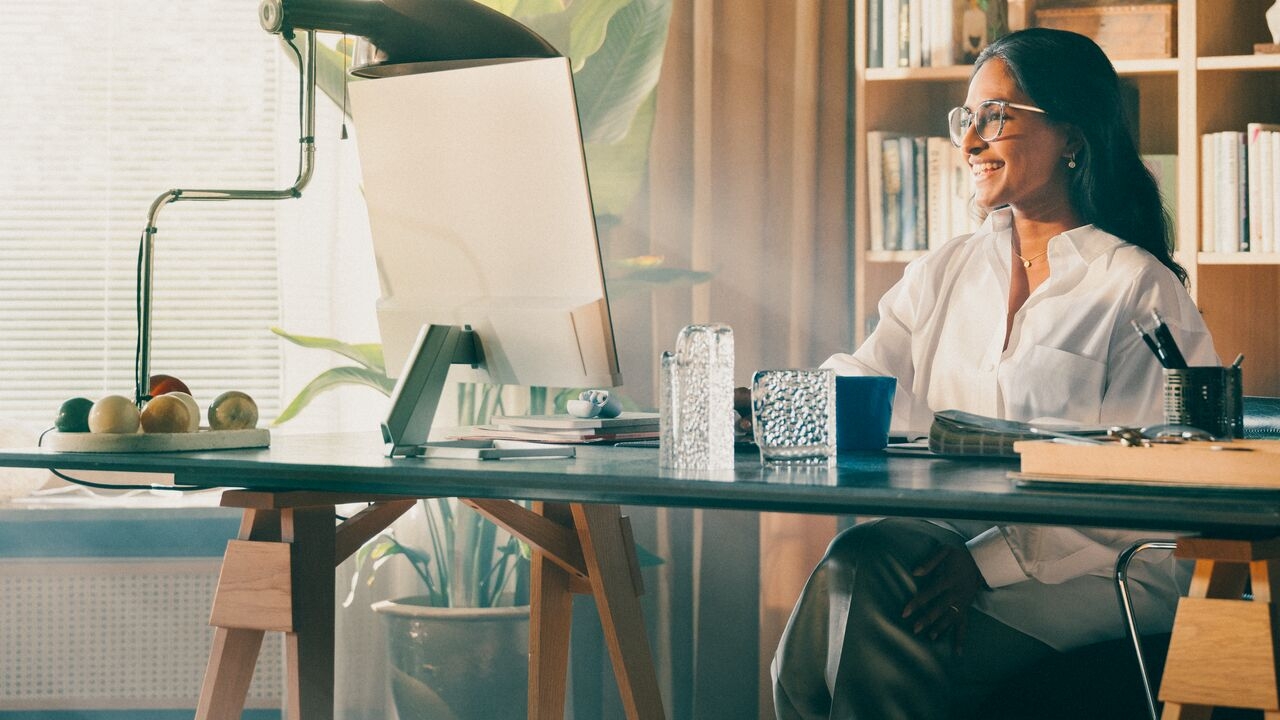 Smiling woman with glasses sitting at a stylish desk with a computer monitor, in a warm, well-decorated home office.