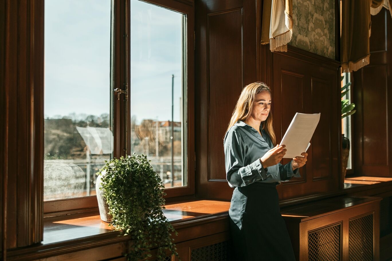 Woman standing by a sunlit window in a classic interior, reading through documents with a plant nearby.
