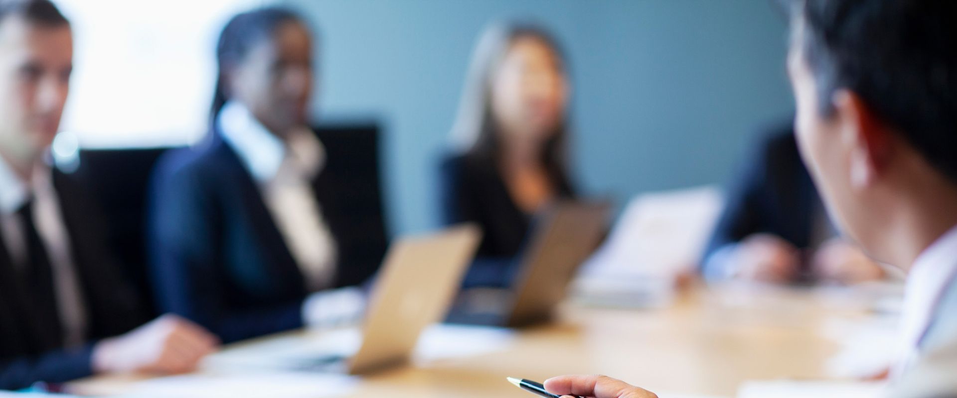 A group of business professionals in formal attire sit around a conference table during a meeting. Several laptops and documents are on the table, and one person in the foreground holds a pen while speaking. The background is softly blurred, keeping the focus on the discussion atmosphere
