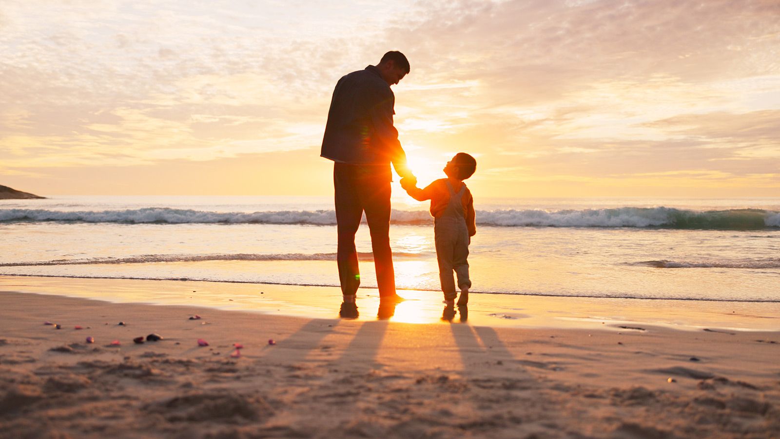 Mand og barn, der holder i hånd på stranden om sommeren som silhouetter i sandet. Familie set bagfra, der går langs havet ved solnedgang.