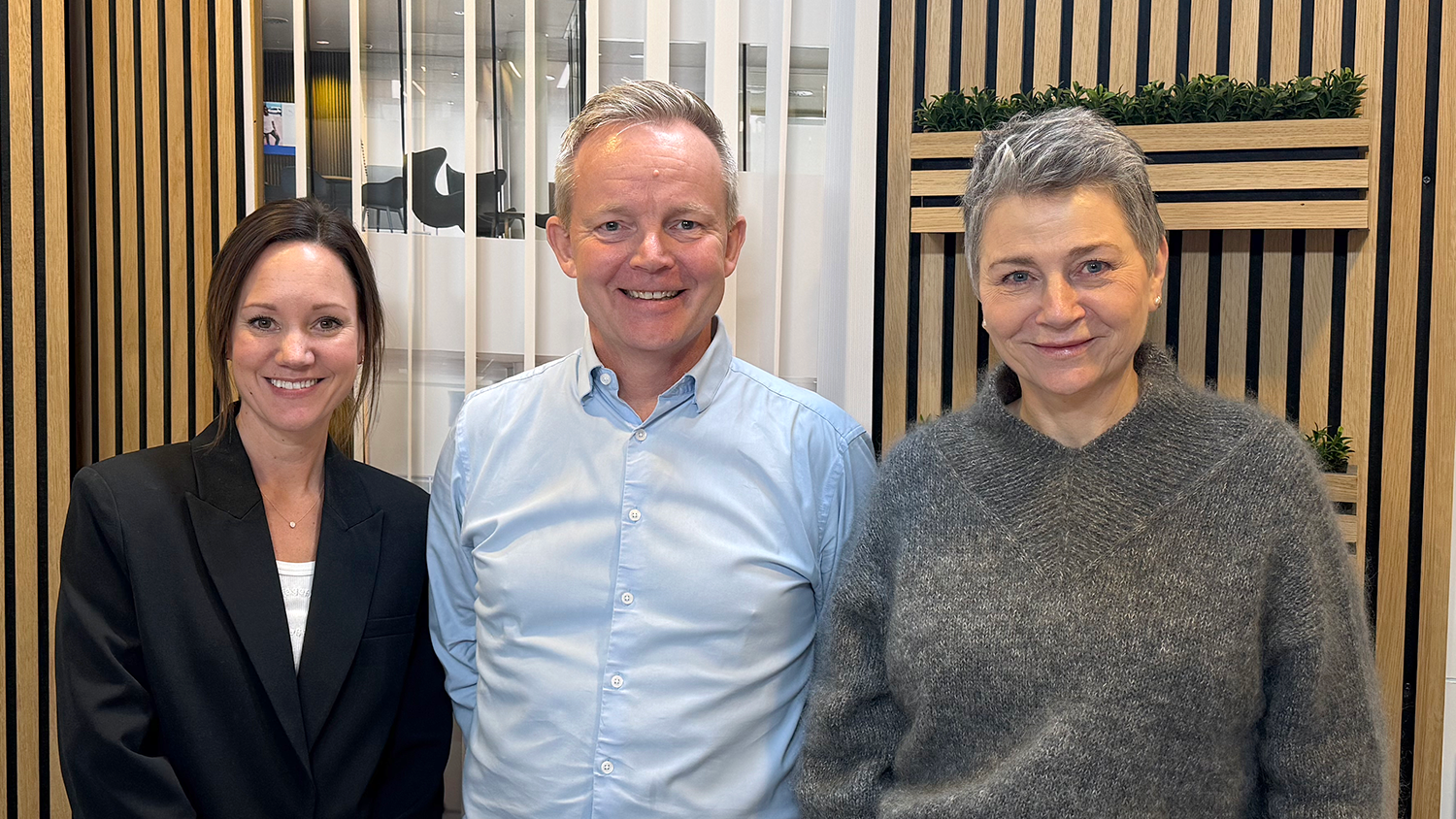 Foto af tre personer, der står side om side indendørs i et moderne kontormiljø med lodrette trælameller og glasvægge i baggrunden. Fra venstre ses privatøkonomisk ekspert Betina Grimstrup, i midten Nordeas investeringsekspert Lars-Kristian Olsen og til højre Pernille Dehn. Alle tre smiler mod kameraet og er klædt i pænt, professionelt tøj.