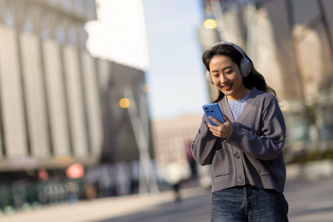 Young woman smiling at her phone while wearing headphones, walking outdoors in a sunny urban setting