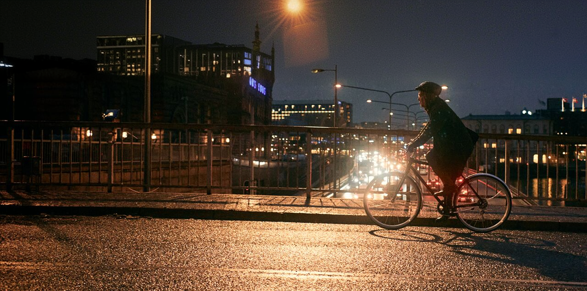 Person cycling across a bridge at night in a city, with street lights and traffic visible in the background.