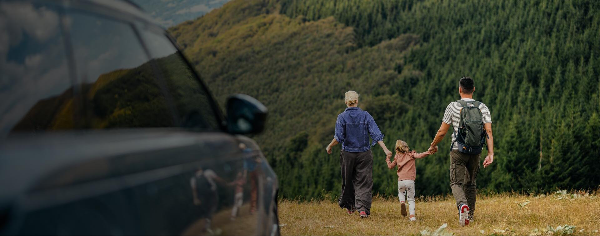 A family of three — two adults and a young child — walking hand in hand through a mountain meadow towards a forest, with a car in the foreground.