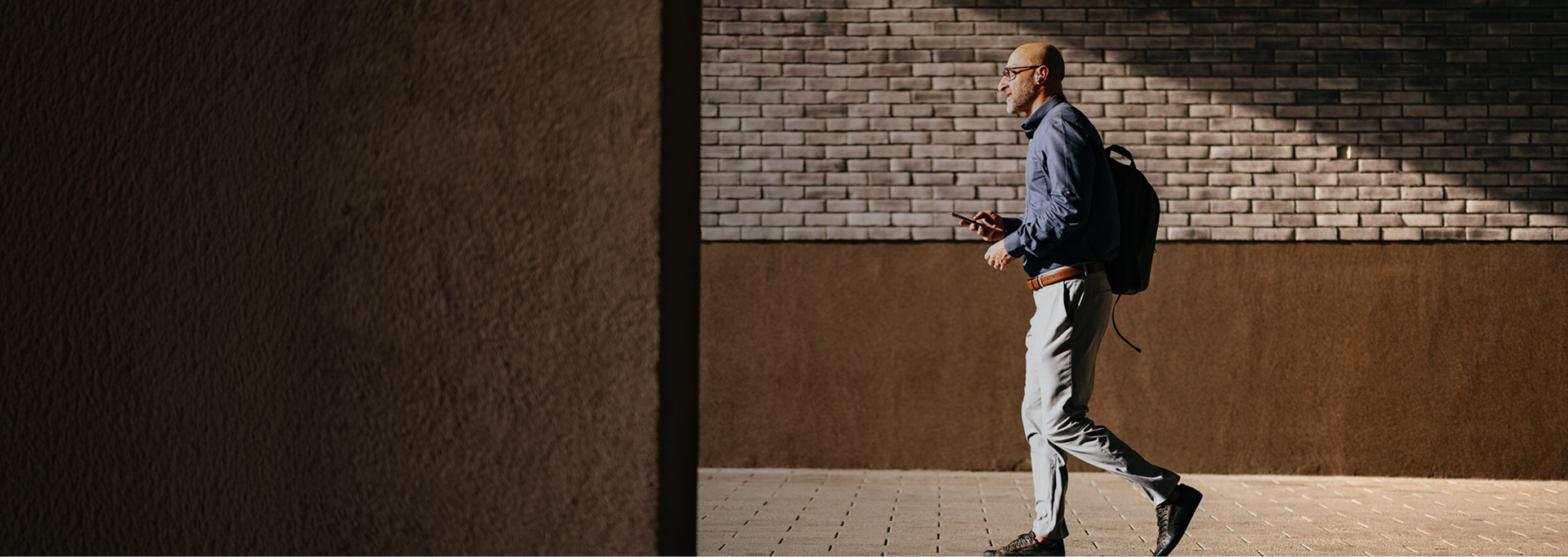 Man walking briskly along a sunlit urban path, holding a phone and wearing a backpack, with a brick wall in the background.