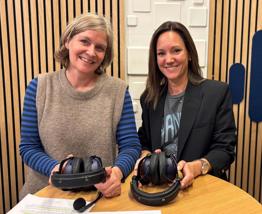 Foto af to kvinder, der sidder ved et bord i et studie og smiler mod kameraet, mens de holder hver deres sæt hovedtelefoner.  