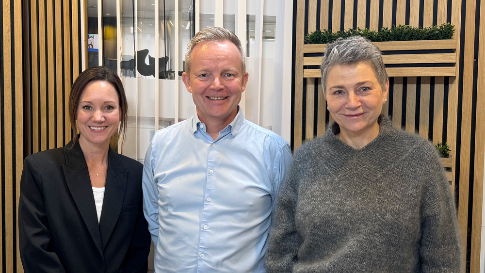 Foto af tre personer, der står side om side indendørs i et moderne kontormiljø med lodrette trælameller og glasvægge i baggrunden. Fra venstre ses privatøkonomisk ekspert Betina Grimstrup, i midten Nordeas investeringsekspert Lars-Kristian Olsen og til højre Pernille Dehn. Alle tre smiler mod kameraet og er klædt i pænt, professionelt tøj.