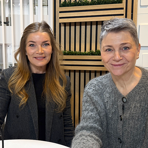 oto af to kvinder, der sidder side om side ved et rundt bord og kigger direkte ind i kameraet. Kvinden til venstre har langt lyst hår og er iført en mørk blazer, og kvinden til højre har kort gråt hår og en grå strikket cardigan. De smiler begge let. I baggrunden ses en indendørs væg med lodrette trælameller og grønne planter.